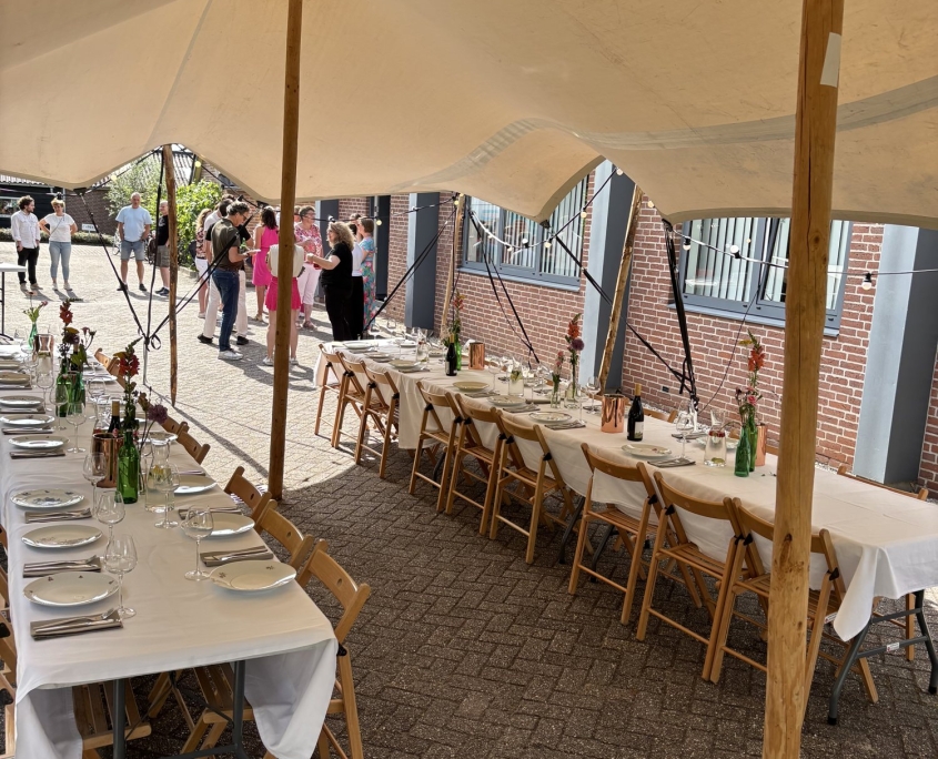 Gedekte lange tafels onder een stretchtent naast een bakstenen loods Gedekte lange tafels onder een stretchtent naast een bakstenen loods