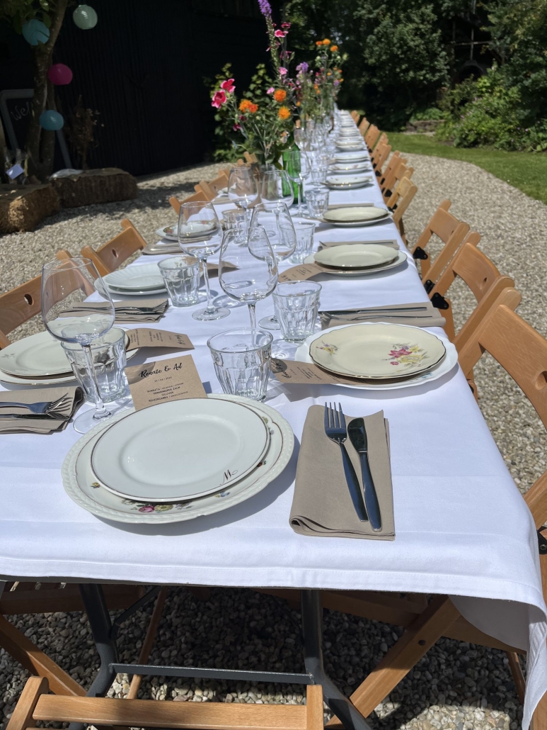 Lange tafel met wit tafelkleed en brocante servies in een tuin Lange tafel met wit tafelkleed en brocante servies in een tuin, Offroad Catering